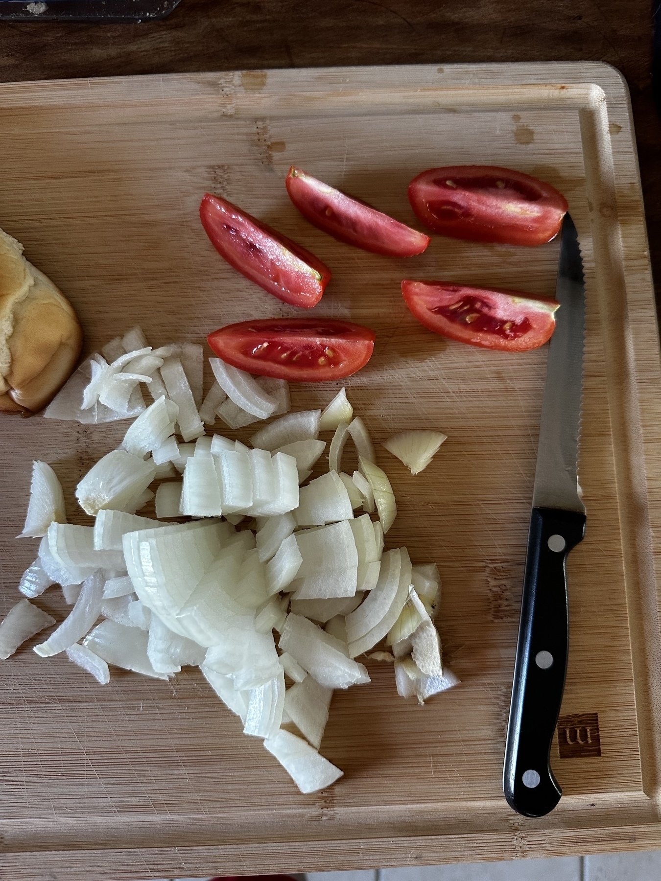 Sliced tomatoes, chopped white onions, a bun, and a knife on a wood chopping board 